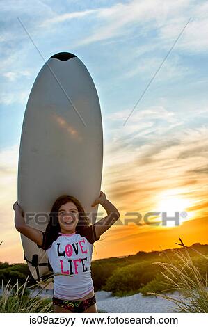 Young girl at beach, carrying surfboard, at sunset View Large Photo Image Stock Photo - Young girl at beach, carrying surfboard, at sunset. Fotosearch