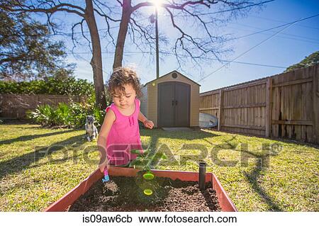 Stock Image - Young girl in garden, planting plant in tub, holding trowel. Fotosearch