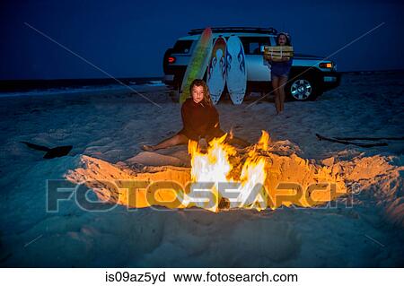 Young girl sitting by campfire on beach, her sister carrying fire wood towards fire View Large Photo Image Stock Image - Young girl sitting by campfire on beach, her sister carrying fire wood towards fire. Fotosearch