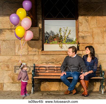 Young girl standing holding bunch of balloons, mature couple sitting on bench watching her View Large Photo Image Stock Image - Young girl standing holding bunch of balloons, mature couple sitting on bench watching her. Fotosearch