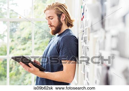 Stock Image - Young male designer leaning against mood board in creative studio looking at digital tablet. Fotosearch