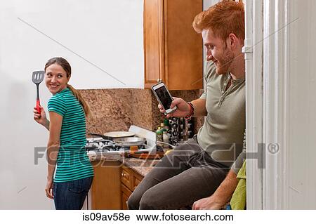 Young man in kitchen using smartphone to take photograph of young woman holding spatula View Large Photo Image Stock Image - Young man in kitchen using smartphone to take photograph of young woman holding spatula. Fotosearch