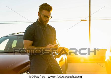 Young man leaning against car, using smartphone View Large Photo Image Stock Photo - Young man leaning against car, using smartphone. Fotosearch