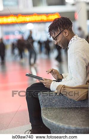 Stock Image - Young man sitting in train station using digital tablet touchscreen. Fotosearch