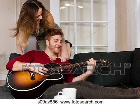 Young man sitting on sofa playing guitar to young woman smiling View Large Photo Image Stock Photograph - Young man sitting on sofa playing guitar to young woman smiling. Fotosearch