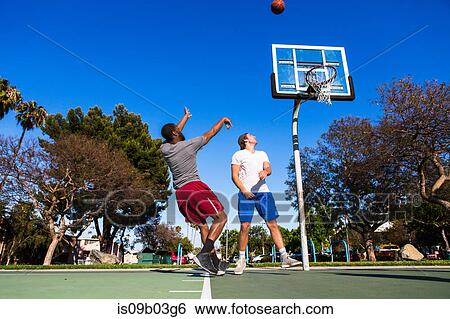 Stock Photograph - Young man throwing basketball towards basketball net on outdoor court. Fotosearch