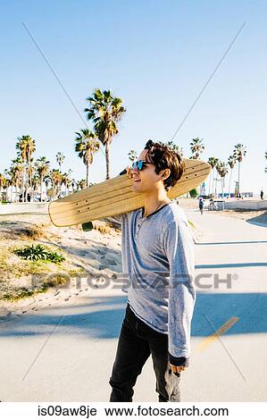 Stock Image - Young man walking outdoors, carrying skateboard on shoulder. Fotosearch