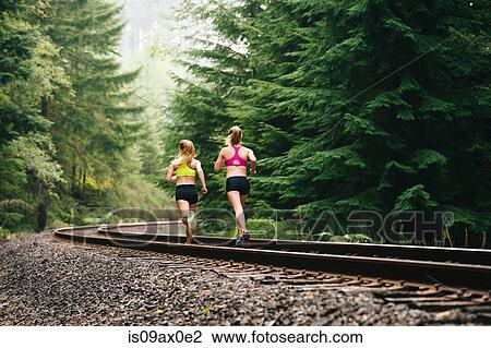 Stock Image - Young woman and teenage girl running along rural train track, rear view. Fotosearch