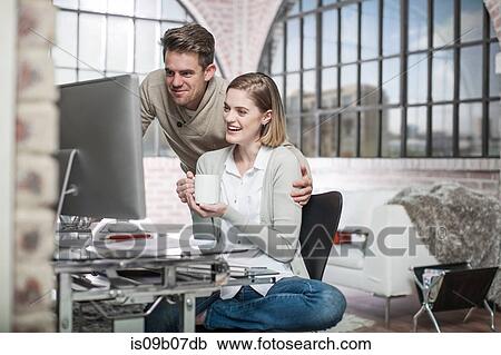 Young woman at home, sitting at desk, looking at computer screen, man beside her, smiling View Large Photo Image Stock Image - Young woman at home, sitting at desk, looking at computer screen, man beside her, smiling. Fotosearch