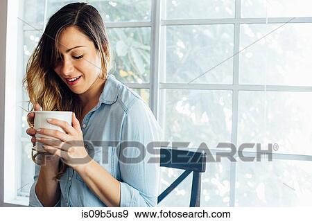 Young woman by window cupping coffee mug View Large Photo Image Stock Photo - Young woman by window cupping coffee mug. Fotosearch