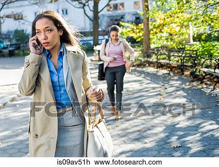 Stock Image - Young woman chatting on smartphone whilst walking through city park. Fotosearch