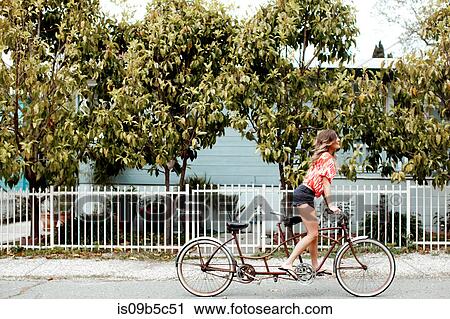 Stock Image - Young woman cycling alone on tandem bicycle along street. Fotosearch