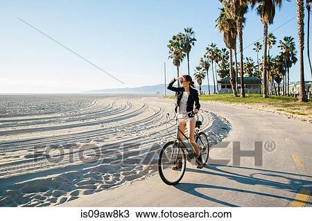 Stock Image - Young woman cycling at beach looking out to sea, Venice Beach, California, USA. Fotosearch