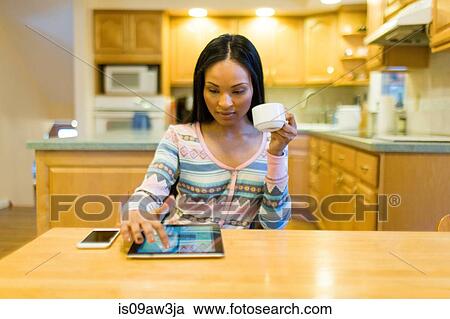 Stock Image - Young woman drinking coffee and using digital tablet touchscreen at kitchen table. Fotosearch