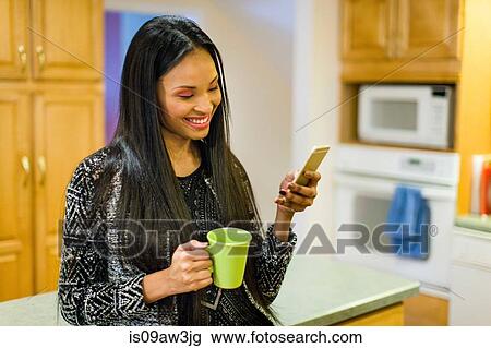 Stock Image - Young woman drinking coffee in kitchen whilst reading smartphone text. Fotosearch