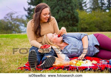 Stock Image - Young woman feeding friend strawberries, in park. Fotosearch