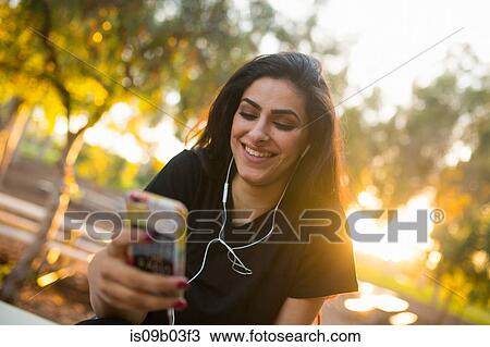 Stock Image - Young woman, outdoors, wearing earphones, holding smartphone. Fotosearch