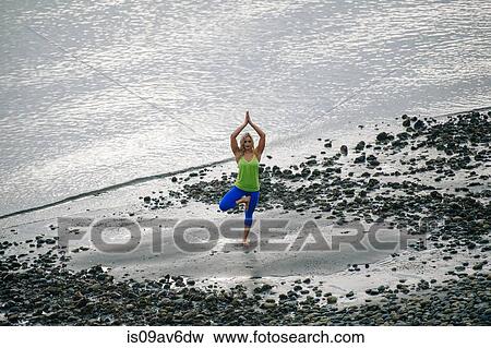 Stock Photo - Young woman practicing yoga standing tree pose on beach. Fotosearch