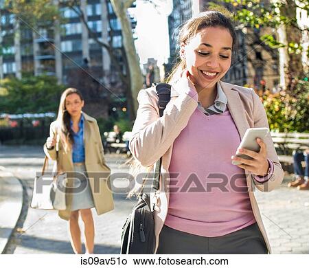 Picture - Young woman reading smartphone texts whilst walking through city park. Fotosearch