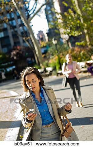 Stock Image - Young woman reading smartphone whilst walking through city park. Fotosearch