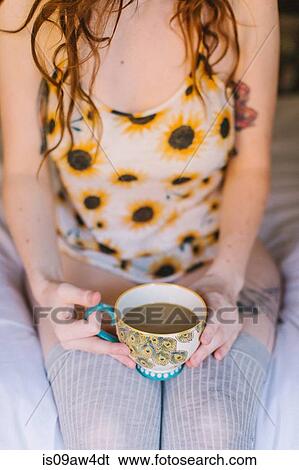 Stock Photograph - Young woman sitting on bed, holding teacup, mid section. Fotosearch