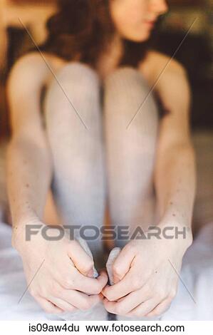 Stock Photograph - Young woman sitting on bed, holding feet, low section. Fotosearch