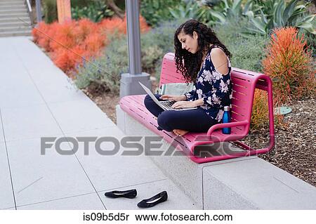 Stock Photography - Young woman sitting on bench, using laptop. Fotosearch