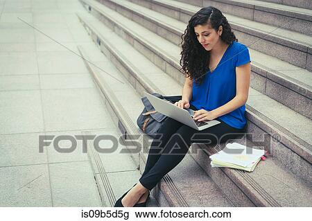 Stock Image - Young woman sitting on steps, using laptop. Fotosearch