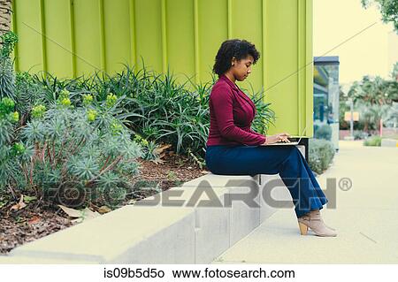 Picture - Young woman sitting on wall, using laptop. Fotosearch