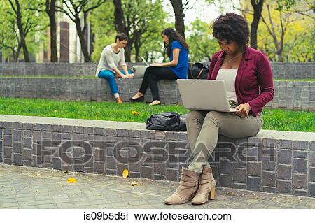 Stock Image - Young woman sitting on wall, using laptop. Fotosearch