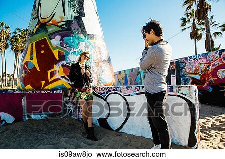 Picture - Young woman standing by graffiti art at beach, young man photographing her. Fotosearch