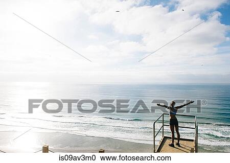 Stock Image - Young woman standing on look out point beside sea, arms raised, rear view. Fotosearch
