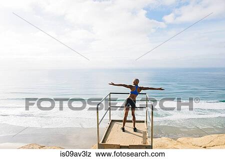 Young woman standing on look out point beside sea, arms raised, rear view View Large Photo Image Stock Photo - Young woman standing on look out point beside sea, arms raised, rear view. Fotosearch