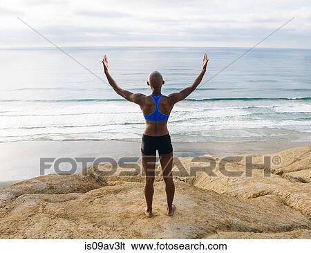 Stock Photograph - Young woman standing on rocks beside sea, arms raised, rear view. Fotosearch