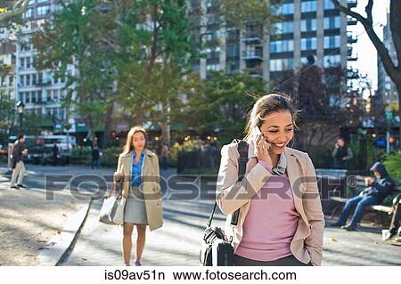 Picture - Young woman talking on smartphone whilst walking through city park. Fotosearch