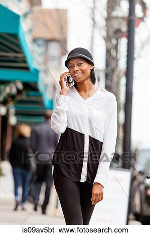 Stock Photograph - Young woman walking along street, using smartphone. Fotosearch
