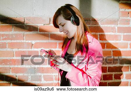 Young woman wearing headphones holding smartphone smiling View Large Photo Image Stock Photograph - Young woman wearing headphones holding smartphone smiling. Fotosearch