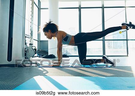 Stock Image - Young woman working out in gym, using gym equipment. Fotosearch