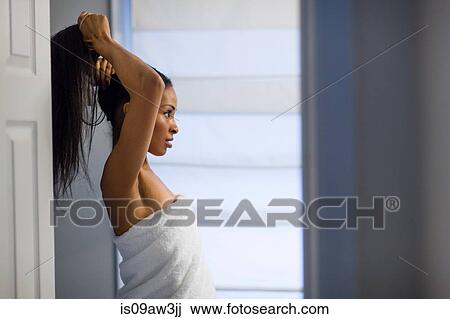 Stock Image - Young woman wrapped in white towel tying up long black hair in bathroom. Fotosearch