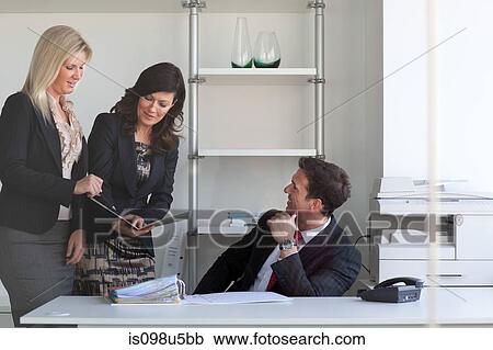 Stock Image - Businesswomen talking to businessman at desk. Fotosearch