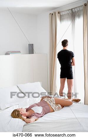 Stock Photograph - Woman lying on bed and man looking out of window. Fotosearch