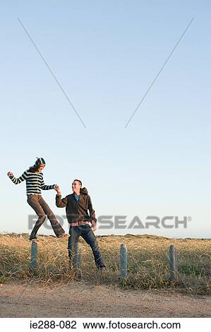 Stock Image - A young man helping a woman balancing on a fence. Fotosearch
