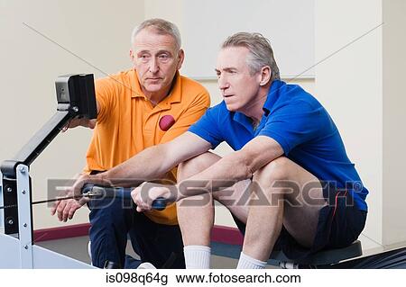 Stock Image - Personal trainer helping a man on a rowing machine. Fotosearch