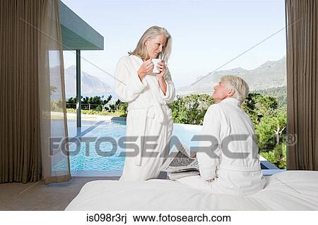 Stock Image - Middle aged couple wearing bathrooms in bedroom with view out to swimming pool. Fotosearch