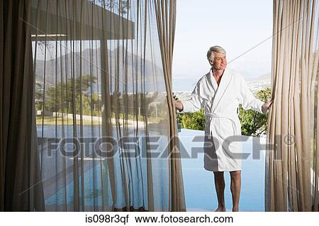 Stock Image - Middle aged man in robe pulling back poolside curtains. Fotosearch