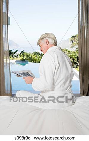 Stock Image - Middle aged man in robe sitting on bed reading newspaper with view out to swimming pool. Fotosearch
