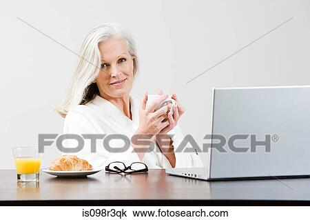 Stock Image - Middle aged woman eating breakfast while using laptop. Fotosearch