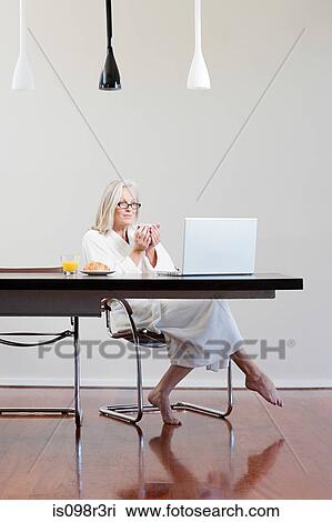 Stock Image - Middle aged woman sitting at table with breakfast and laptop. Fotosearch
