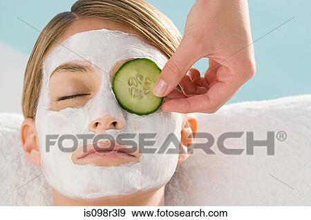 Stock Photo - Young woman wearing facial mask having cucumber slice placed over eye. Fotosearch