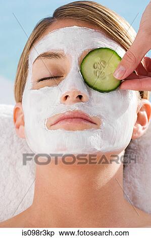 Stock Photography - Young woman with facial mask having cucumber slice placed over eye. Fotosearch
